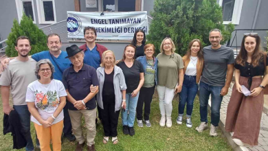 Görselin üst bölümünde, bir binanın bahçesindeki yeşil çim alanda yan yana dizilerek objektife gülümseyen kalabalık bir sağlık ekibi ve katılımcı grubu yer alıyor. Arka plandaki binanın giriş kapısı önünde "ENGEL TANIMAYAN DİŞ HEKİMLİĞİ DERNEĞİ - İZMİR" yazılı ve dernek amblemli beyaz bir pankart asılı duruyor. Ön sırada siyah şapkalı, lacivert polo tişörtlü yaşlı bir beyefendi ile yanında açık gri hırkalı kadın, önünde ise turuncu pantolonlu ve desenli beyaz tişörtlü bir kadın yer alıyor. Grubun geri kalanında ise diş hekimi formaları (mavi ameliyathane forması), tişört, kot pantolon ve uzun etek gibi günlük kıyafetler giymiş kadın ve erkek hekimler ile gönüllüler birbirlerine sarılarak poz veriyor; arkada ise yemyeşil çam ağaçları ve bina cephesi görülüyor.