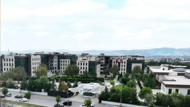 Görselin üst bölümünde, Bursa Teknik Üniversitesi (BTÜ) kampüs yerleşkesinin geniş açılı havadan (kuş bakışı) görünümü yer alıyor. Ön planda araçların geçtiği çok şeritli asfalt bir cadde ve üniversitenin modern nizamiye/güvenlik giriş kapısı görülürken; orta alanda yeşil ağaçlarla çevrili peyzaj düzenlemesi, otopark alanları ve üzerinde "REKTÖRLÜK" tabelası ile Türk bayrağı asılı çok katlı modern fakülte binaları uzanıyor. Arka planda ise hafif bulutlu açık gökyüzü altında şehri çevreleyen dağ sırası ve vadi manzarası seçiliyor.