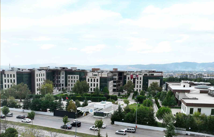 Görselin üst bölümünde, Bursa Teknik Üniversitesi (BTÜ) kampüs yerleşkesinin geniş açılı havadan (kuş bakışı) görünümü yer alıyor. Ön planda araçların geçtiği çok şeritli asfalt bir cadde ve üniversitenin modern nizamiye/güvenlik giriş kapısı görülürken; orta alanda yeşil ağaçlarla çevrili peyzaj düzenlemesi, otopark alanları ve üzerinde "REKTÖRLÜK" tabelası ile Türk bayrağı asılı çok katlı modern fakülte binaları uzanıyor. Arka planda ise hafif bulutlu açık gökyüzü altında şehri çevreleyen dağ sırası ve vadi manzarası seçiliyor.