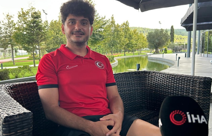 Görselin üst bölümünde, açık alandaki bir tesiste hasır koltukta oturarak röportaj veren milli ampute futbolcu Barış Telli yer alıyor. Kıvırcık koyu renk saçlı ve hafif sakallı olan Telli, göğsünde Türk bayrağı arması bulunan kırmızı milli takım polo tişörtü ve siyah şort giymiş halde tebessüm ederek kameraya bakıyor. Sağ alt köşede İhlas Haber Ajansı (İHA) logolu mikrofon ucu görülürken; arka planda ise yeşil ağaçlar, çimlik alan, yapay gölet/havuz ve modern tesis yapıları seçiliyor.