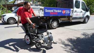 Görselde, kırmızı tişörtlü bir görevli akülü tekerlekli sandalyeyi yol üzerinde ilerletiyor. Arka planda üzerinde Sosyal Yardım Merkezi yazısı bulunan belediyeye ait bir araç ve başka bir görevli görülüyor.