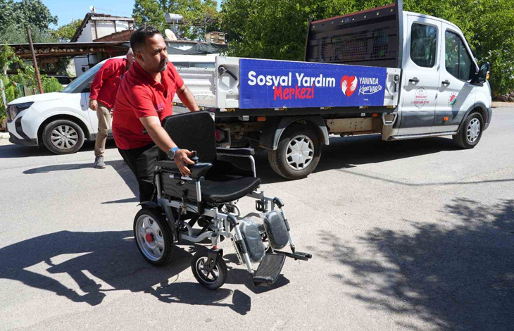 Görselde, kırmızı tişörtlü bir görevli akülü tekerlekli sandalyeyi yol üzerinde ilerletiyor. Arka planda üzerinde Sosyal Yardım Merkezi yazısı bulunan belediyeye ait bir araç ve başka bir görevli görülüyor.
