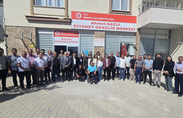rselin üst bölümünde, T.C. Besni İlçe Müftülüğü Ahmet Saçlı Diyanet Gençlik Merkezi binasının önünde toplanarak toplu hatıra fotoğrafı çektiren kalabalık bir heyet ve vatandaş grubu yer alıyor. Binanın girişinde kırmızı-beyaz tabelada merkez adı okunurken, ön cephedeki camların önünde takım elbiseli protokol üyeleri, kadın ve erkek katılımcılar geniş bir sıra halinde dizilmiş durumda. Topluluğun tam ortasında mavi tişörtlü bedensel engelli bir birey tekerlekli sandalyesinde oturarak objektife bakarken, iki yanında ve arkasında yerel yetkililer, din görevlileri ve sivil katılımcılar kilit parke taşlı avluda yan yana durarak gülümsüyor.