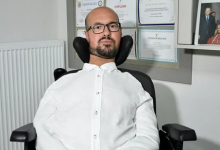 Emre Vural, tekerlekli sandalyede otururken görülüyor. Beyaz gömlek ve gözlük takıyor. Arka planda duvarda çeşitli sertifikalar ve bir fotoğraf çerçevesi yer alıyor; ortam profesyonel bir çalışma alanını andırıyor.