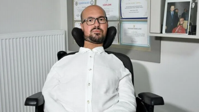 Emre Vural, tekerlekli sandalyede otururken görülüyor. Beyaz gömlek ve gözlük takıyor. Arka planda duvarda çeşitli sertifikalar ve bir fotoğraf çerçevesi yer alıyor; ortam profesyonel bir çalışma alanını andırıyor.