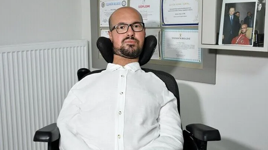 Emre Vural, tekerlekli sandalyede otururken görülüyor. Beyaz gömlek ve gözlük takıyor. Arka planda duvarda çeşitli sertifikalar ve bir fotoğraf çerçevesi yer alıyor; ortam profesyonel bir çalışma alanını andırıyor.