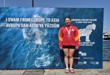 Görselde, kırmızı Galatasaray eşofman üstü ve siyah şort giyen, boynunda madalya bulunan Selin Zülal Önal, Çanakkale Boğazı’nı yüzerek geçmesinin ardından “I SWAM FROM EUROPE TO ASIA” yazılı etkinlik panosunun önünde gururla poz veriyor.