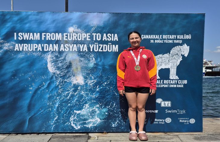 Görselde, kırmızı Galatasaray eşofman üstü ve siyah şort giyen, boynunda madalya bulunan Selin Zülal Önal, Çanakkale Boğazı’nı yüzerek geçmesinin ardından “I SWAM FROM EUROPE TO ASIA” yazılı etkinlik panosunun önünde gururla poz veriyor.