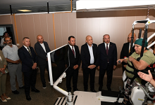 Solda açılışa katılan protokol üyeleri bir arada duruyor ve cihazın tanıtımını izliyor. Sağ tarafta ise bir kadın, robotik yürüme cihazına bağlı şekilde yürümeyi deneyimliyor. Ortam, bir hastanenin fizik tedavi veya rehabilitasyon birimini andırıyor.