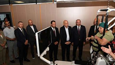 Solda açılışa katılan protokol üyeleri bir arada duruyor ve cihazın tanıtımını izliyor. Sağ tarafta ise bir kadın, robotik yürüme cihazına bağlı şekilde yürümeyi deneyimliyor. Ortam, bir hastanenin fizik tedavi veya rehabilitasyon birimini andırıyor.