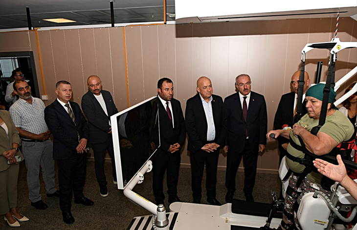 Solda açılışa katılan protokol üyeleri bir arada duruyor ve cihazın tanıtımını izliyor. Sağ tarafta ise bir kadın, robotik yürüme cihazına bağlı şekilde yürümeyi deneyimliyor. Ortam, bir hastanenin fizik tedavi veya rehabilitasyon birimini andırıyor.