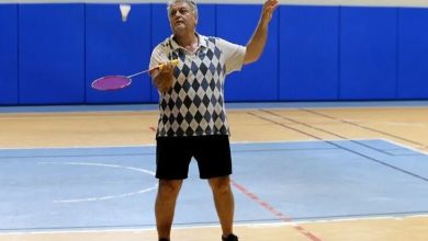 Kapalı bir spor salonunda, badminton sahasında Cavit Şengün görülüyor. Üzerinde kısa kollu, baklava desenli bir spor tişört, siyah şort ve spor ayakkabıları var. Elinde badminton raketiyle havada duran tüy topa doğru hamle yapmak üzere. Duruşu dinamik, odaklanmış ve oyuna hâkim bir sporcu izlenimi veriyor. Arka planda mavi minderlerle kaplı duvarlar ve zeminde kırmızı‑mavi saha çizgileri bulunuyor. Ortam aydınlık, düzenli ve tipik bir salon spor alanı atmosferi taşıyor.