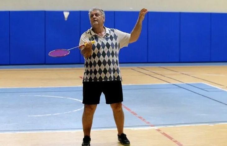 Kapalı bir spor salonunda, badminton sahasında Cavit Şengün görülüyor. Üzerinde kısa kollu, baklava desenli bir spor tişört, siyah şort ve spor ayakkabıları var. Elinde badminton raketiyle havada duran tüy topa doğru hamle yapmak üzere. Duruşu dinamik, odaklanmış ve oyuna hâkim bir sporcu izlenimi veriyor. Arka planda mavi minderlerle kaplı duvarlar ve zeminde kırmızı‑mavi saha çizgileri bulunuyor. Ortam aydınlık, düzenli ve tipik bir salon spor alanı atmosferi taşıyor.