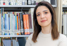 Çocuk ve ergenlerle çalışan Klinik Psikolog Eliz Avcı, sakin ve güven veren bir duruşa sahip. Omuz hizasında düz, açık renk saçları var; açık tonlarda, sade bir üst giymiş. Arkasında farklı renk ve boyutlarda kitaplarla dolu bir raf bulunuyor. Ortam, akademik bir çalışma alanı hissi veriyor. Genel görünümü, profesyonel, dikkatli ve danışanlarına karşı sıcak bir yaklaşımı olan bir uzman izlenimi oluşturuyor.