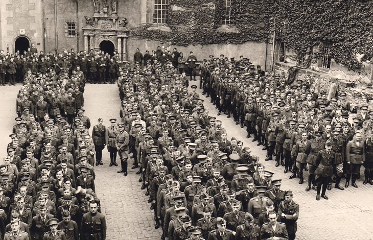 Taş duvarlı, tarihi bir avluda çok sayıda asker düzenli sıralar hâlinde tören için toplanmış görünüyor. Önde birkaç subay duruyor, ortam resmî bir askerî diziliş havasında.