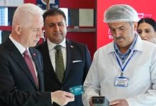 Fotoğrafta İstanbul Valisi Davut Gül, okul kantininde “Gönülden Bir Öğün” projesi kapsamında öğrencilere verilen kantin kartını deniyor. Vali Gül takım elbisesiyle, elindeki kartı kantin görevlisinin tuttuğu POS cihazına yaklaştırırken görülüyor. Kantin görevlisi beyaz önlük ve bone takıyor, üzerinde “KANTİN GÖREVLİSİ” yazılı isimlik bulunuyor. Arka planda kırmızı duvar ve raflar yer alıyor; sahne, projenin uygulamalı tanıtım anını yansıtıyor. Atmosfer resmi, düzenli ve sosyal destek temalı.