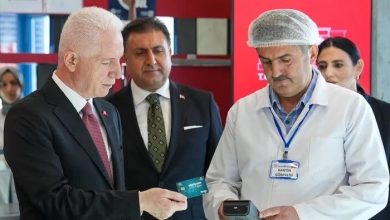 Fotoğrafta İstanbul Valisi Davut Gül, okul kantininde “Gönülden Bir Öğün” projesi kapsamında öğrencilere verilen kantin kartını deniyor. Vali Gül takım elbisesiyle, elindeki kartı kantin görevlisinin tuttuğu POS cihazına yaklaştırırken görülüyor. Kantin görevlisi beyaz önlük ve bone takıyor, üzerinde “KANTİN GÖREVLİSİ” yazılı isimlik bulunuyor. Arka planda kırmızı duvar ve raflar yer alıyor; sahne, projenin uygulamalı tanıtım anını yansıtıyor. Atmosfer resmi, düzenli ve sosyal destek temalı.