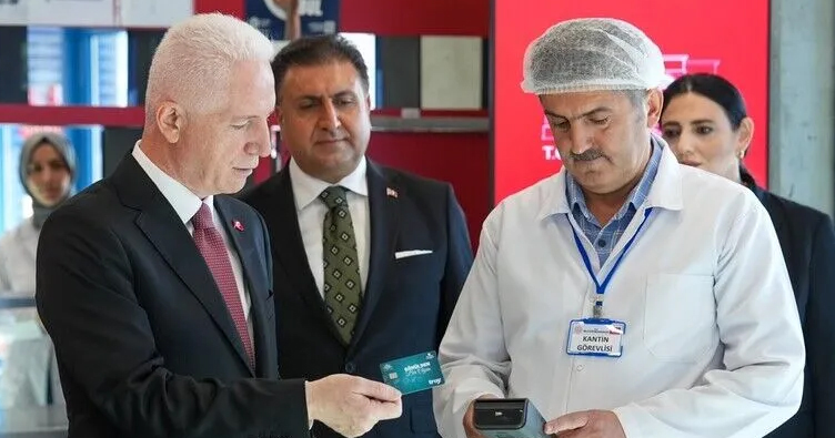 Fotoğrafta İstanbul Valisi Davut Gül, okul kantininde “Gönülden Bir Öğün” projesi kapsamında öğrencilere verilen kantin kartını deniyor. Vali Gül takım elbisesiyle, elindeki kartı kantin görevlisinin tuttuğu POS cihazına yaklaştırırken görülüyor. Kantin görevlisi beyaz önlük ve bone takıyor, üzerinde “KANTİN GÖREVLİSİ” yazılı isimlik bulunuyor. Arka planda kırmızı duvar ve raflar yer alıyor; sahne, projenin uygulamalı tanıtım anını yansıtıyor. Atmosfer resmi, düzenli ve sosyal destek temalı.