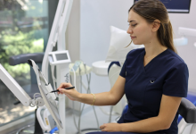 Diş Hekimi İpek Rizvan, lacivert hekim formasıyla klinik ortamında çalışan bir sağlık profesyoneli olarak görülüyor. Dokunmatik ekrana kalemle işlem yaparken arka planda diş üniti, tıbbi ekipmanlar ve geniş bir pencereden gelen doğal ışık yer alıyor. Ortam temiz, modern ve profesyonel bir klinik izlenimi veriyor.