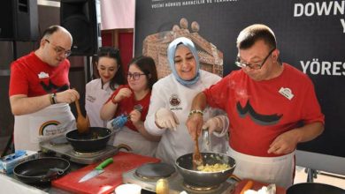 Beş kişi bir masa etrafında birlikte yemek pişiriyor. Herkes önlük takıyor; bazıları eldiven kullanıyor. Masanın üzerinde taşınabilir ocaklar, tavalar, spatulalar ve çeşitli malzemeler bulunuyor. İki kişi tavada yemeği karıştırıyor, diğerleri yardımcı oluyor veya izliyor. Arkada Türkçe bir afiş var; üzerinde “Sempozyumu”, “Vecihi Hürkuş Havacılık ve Teknoloji Parkı” ve “Down” kelimeleri yer alıyor. Ortam, kapsayıcı bir etkinlikte yapılan toplu bir yemek atölyesini gösteriyor.