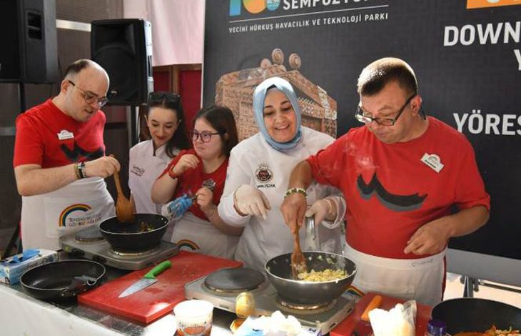 Beş kişi bir masa etrafında birlikte yemek pişiriyor. Herkes önlük takıyor; bazıları eldiven kullanıyor. Masanın üzerinde taşınabilir ocaklar, tavalar, spatulalar ve çeşitli malzemeler bulunuyor. İki kişi tavada yemeği karıştırıyor, diğerleri yardımcı oluyor veya izliyor. Arkada Türkçe bir afiş var; üzerinde “Sempozyumu”, “Vecihi Hürkuş Havacılık ve Teknoloji Parkı” ve “Down” kelimeleri yer alıyor. Ortam, kapsayıcı bir etkinlikte yapılan toplu bir yemek atölyesini gösteriyor.