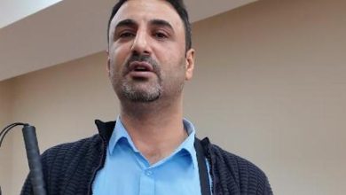 Görme engelli Dr. Önder İşlek, kapalı bir mekânda ayakta duran, kısa saçlı ve hafif sakallı bir yetişkin erkek olarak görülüyor. Açık mavi gömlek ve koyu renk dokulu bir ceket giyiyor. Sağ elinde baston tutuyor; duruşu sakin ve kendinden emin. Arka planda sade, bej tonlarında bir duvar bulunuyor.