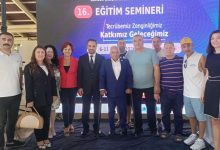Kapalı bir alanda, büyük bir dijital ekranın önünde yan yana duran on kişilik bir grup görülüyor. Ekranda bir eğitim seminerine ait başlık ve tarih bilgileri yer alıyor. Grup, hatıra fotoğrafı çeker gibi kameraya doğru bakıyor. Kimi daha resmi, kimi daha günlük kıyafetler giymiş. Ortam düzenli, aydınlık ve bir etkinlik alanı havasında.