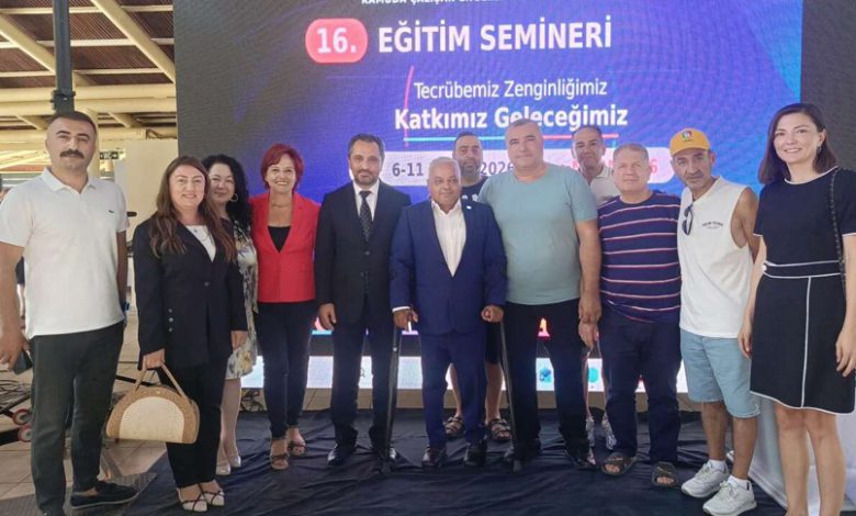 Kapalı bir alanda, büyük bir dijital ekranın önünde yan yana duran on kişilik bir grup görülüyor. Ekranda bir eğitim seminerine ait başlık ve tarih bilgileri yer alıyor. Grup, hatıra fotoğrafı çeker gibi kameraya doğru bakıyor. Kimi daha resmi, kimi daha günlük kıyafetler giymiş. Ortam düzenli, aydınlık ve bir etkinlik alanı havasında.