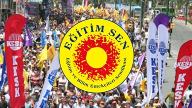 Kalabalık bir yürüyüş ya da gösteri görülüyor. İnsanlar şehir içindeki geniş bir caddede birlikte ilerliyor. Önde büyük, sarı-kırmızı renkli yuvarlak bir pankart taşınıyor; üzerinde “Eğitim Sen” yazısı bulunuyor. Pankartın çevresinde uzun, dikey bayraklar var ve üzerlerinde “KESK” ifadesi yer alıyor. Katılımcılar ellerinde çeşitli dövizler ve flamalar taşıyor. Sahne, örgütlü bir toplu yürüyüşün canlı ve hareketli atmosferini yansıtıyor.