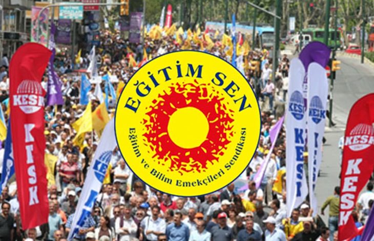 Kalabalık bir yürüyüş ya da gösteri görülüyor. İnsanlar şehir içindeki geniş bir caddede birlikte ilerliyor. Önde büyük, sarı-kırmızı renkli yuvarlak bir pankart taşınıyor; üzerinde “Eğitim Sen” yazısı bulunuyor. Pankartın çevresinde uzun, dikey bayraklar var ve üzerlerinde “KESK” ifadesi yer alıyor. Katılımcılar ellerinde çeşitli dövizler ve flamalar taşıyor. Sahne, örgütlü bir toplu yürüyüşün canlı ve hareketli atmosferini yansıtıyor.