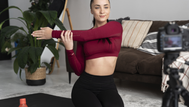 Bir kişi ev ortamında yoga matı üzerinde diz çökmüş pozisyonda, kolunu göğsünün önünde esnetiyor. Önünde tripod üzerinde bir kamera bulunuyor; bu da kişinin egzersiz kaydı veya çevrim içi içerik hazırladığını gösteriyor. Matın üzerinde bir su şişesi var. Arka planda koltuk, bitki ve merdiven görülen sıcak bir salon ortamı yer alıyor.