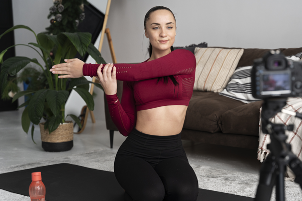 Bir kişi ev ortamında yoga matı üzerinde diz çökmüş pozisyonda, kolunu göğsünün önünde esnetiyor. Önünde tripod üzerinde bir kamera bulunuyor; bu da kişinin egzersiz kaydı veya çevrim içi içerik hazırladığını gösteriyor. Matın üzerinde bir su şişesi var. Arka planda koltuk, bitki ve merdiven görülen sıcak bir salon ortamı yer alıyor.