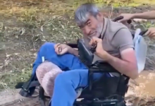 Görselin üst bölümünde, açık alanda akülü tekerlekli sandalyesinde oturan bedensel engelli bir adam yer alıyor. Kır saçlı ve sakallı olan adam, bej tonlarında polo yaka bir tişört ile mavi eşofman altı giymiş vaziyette tebessümle kameraya bakıyor. Sandalyesinin itme kollarından tutan refakatçilerin elleri ile arka planda toprak zemin ve seyrek yeşil bitki örtüsü görülüyor.