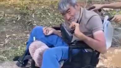 Görselin üst bölümünde, açık alanda akülü tekerlekli sandalyesinde oturan bedensel engelli bir adam yer alıyor. Kır saçlı ve sakallı olan adam, bej tonlarında polo yaka bir tişört ile mavi eşofman altı giymiş vaziyette tebessümle kameraya bakıyor. Sandalyesinin itme kollarından tutan refakatçilerin elleri ile arka planda toprak zemin ve seyrek yeşil bitki örtüsü görülüyor.