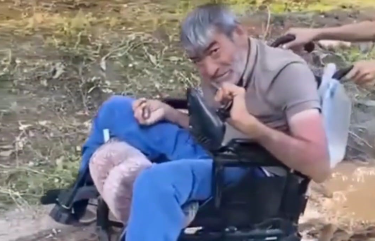 Görselin üst bölümünde, açık alanda akülü tekerlekli sandalyesinde oturan bedensel engelli bir adam yer alıyor. Kır saçlı ve sakallı olan adam, bej tonlarında polo yaka bir tişört ile mavi eşofman altı giymiş vaziyette tebessümle kameraya bakıyor. Sandalyesinin itme kollarından tutan refakatçilerin elleri ile arka planda toprak zemin ve seyrek yeşil bitki örtüsü görülüyor.