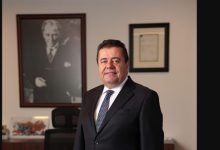 Fotoğrafta Ender Güzeler, resmi ve kurumsal bir ofis ortamında ayakta duruyor. Üzerinde koyu renk bir takım elbise, beyaz gömlek ve desenli bir kravat bulunuyor. Duruşu kendinden emin, profesyonel ve sakin. Arka planda duvarda büyük boy siyah‑beyaz bir portre ile çerçeveli bir belge yer alıyor. Yan tarafta ofis koltuğu ve masada bazı eşyalar görülüyor. Genel atmosfer, kurumsal bir yöneticinin çalışma alanını yansıtan ciddi ve düzenli bir ortam hissi veriyor.