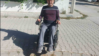 Görselin üst bölümünde, kilit parke taşlarıyla döşenmiş ve yer yer taşları sökülüp bozulmuş bir sokakta siyah akülü tekerlekli sandalyesiyle duran yetişkin bir erkek yer alıyor. Çizgili koyu renkli uzun kollu tişört, gri pantolon ve spor ayakkabı giymiş olan birey, bir eli kumanda kolunda olacak şekilde doğrudan objektife bakıyor. Hemen önünde zemindeki çökmüş ve yerinden oynamış parke taşları görülürken, arka planda beyaz boyalı bir bahçe duvarı, yeşil sarmaşık yaprakları, elektrik direği ve yol kenarındaki çöp konteyneri seçiliyor.