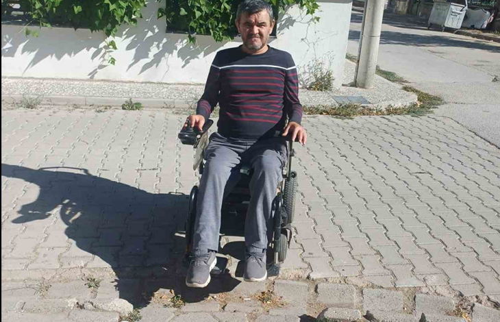 Görselin üst bölümünde, kilit parke taşlarıyla döşenmiş ve yer yer taşları sökülüp bozulmuş bir sokakta siyah akülü tekerlekli sandalyesiyle duran yetişkin bir erkek yer alıyor. Çizgili koyu renkli uzun kollu tişört, gri pantolon ve spor ayakkabı giymiş olan birey, bir eli kumanda kolunda olacak şekilde doğrudan objektife bakıyor. Hemen önünde zemindeki çökmüş ve yerinden oynamış parke taşları görülürken, arka planda beyaz boyalı bir bahçe duvarı, yeşil sarmaşık yaprakları, elektrik direği ve yol kenarındaki çöp konteyneri seçiliyor.