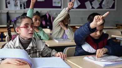 Görselin üst bölümünde, bir okul sınıfında sıralarda oturan ve derse katılmak için parmak kaldıran görme engelli öğrenciler yer alıyor. Ön planda sol tarafta, kırmızı çerçeveli gözlük takan, saçları örgülü ve ekose gömlekli bir kız çocuğu önündeki kabartma Braille alfabesiyle basılmış açık kitaba dokunarak dikkatle konuşuyor. Yanında oturan montlu erkek öğrenci sağ elini havaya kaldırıp diğer eliyle sıradaki kabartmalı çalışma kâğıdını takip ederken; arka sıralarda oturan diğer öğrenciler de derse katılmak amacıyla heyecanla parmak kaldırıyor.