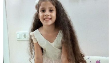 Dolunay Elçi kameraya dönük ve gülümsüyor. Uzun, koyu kahverengi ve dalgalı saçları omuzlarının ve gövdesinin iki yanına doğru uzanıyor; saçlarının bir kısmı geriye alınmış. Üzerinde açık krem/beyaz tonlarında, kısa kollu ve dantel detaylı bir elbise var.
