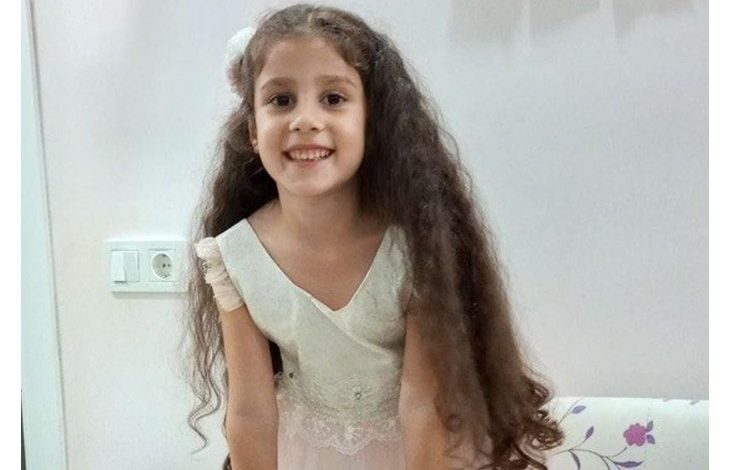 Dolunay Elçi kameraya dönük ve gülümsüyor. Uzun, koyu kahverengi ve dalgalı saçları omuzlarının ve gövdesinin iki yanına doğru uzanıyor; saçlarının bir kısmı geriye alınmış. Üzerinde açık krem/beyaz tonlarında, kısa kollu ve dantel detaylı bir elbise var.