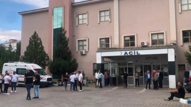 Görselin üst bölümünde, bir devlet hastanesinin acil servis binası ve önündeki açık alan yer alıyor. Pembe-bej tonlarındaki çok katlı hastane binasının zemin katında üzerinde büyük harflerle "ACİL" yazılı giriş kapısı ve sundurması bulunuyor. Sol tarafta park halinde beyaz bir 112 Acil Sağlık ambulansı dururken, hastane bahçesindeki kilit taşlı zeminde bekleyen hasta yakınları, vatandaşlar ve sağlık personeli gruplar halinde görülüyor. Sağ alt köşede ise kaldırım kenarına oturarak bekleyen bir kişi yer alıyor.