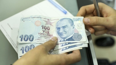 Görselde, bir kişinin elinde birkaç adet 100 Türk lirası banknot tuttuğu ve diğer eliyle kalemle bir belge üzerinde işlem yaptığı görülüyor. Arka planda evraklar ve masa üzerindeki ofis ekipmanları yer alıyor.