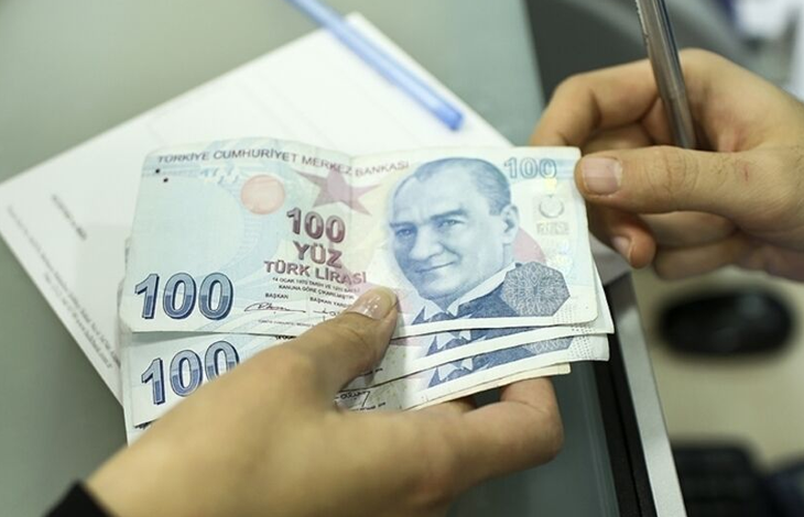 Görselde, bir kişinin elinde birkaç adet 100 Türk lirası banknot tuttuğu ve diğer eliyle kalemle bir belge üzerinde işlem yaptığı görülüyor. Arka planda evraklar ve masa üzerindeki ofis ekipmanları yer alıyor.