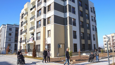 Modern bir sosyal konut yerleşimi görülüyor. Çok katlı, açık renk cepheli apartman bloklarının önünde geniş ve düzenli bir yaya alanı bulunuyor. Bu alanda tekerlekli sandalye kullanan bireyler, beyaz baston ve rehber köpek eşliğinde yürüyen bir kişi gibi farklı erişilebilirlik ihtiyaçlarına sahip insanlar rahatça hareket ediyor. Binaların girişleri düz zeminle bağlantılı, yollar geniş, peyzaj düzeni temiz ve engelsiz. Ortam, erişilebilirlik ilkelerine uygun tasarlanmış bir sosyal konut projesinin kapsayıcı yapısını yansıtıyor; herkesin bağımsız ve güvenli biçimde dolaşabildiği, eşit kullanım imkânı sunan bir yaşam alanı hissi veriyor.
