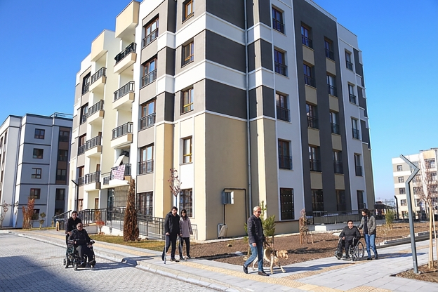 Modern bir sosyal konut yerleşimi görülüyor. Çok katlı, açık renk cepheli apartman bloklarının önünde geniş ve düzenli bir yaya alanı bulunuyor. Bu alanda tekerlekli sandalye kullanan bireyler, beyaz baston ve rehber köpek eşliğinde yürüyen bir kişi gibi farklı erişilebilirlik ihtiyaçlarına sahip insanlar rahatça hareket ediyor. Binaların girişleri düz zeminle bağlantılı, yollar geniş, peyzaj düzeni temiz ve engelsiz. Ortam, erişilebilirlik ilkelerine uygun tasarlanmış bir sosyal konut projesinin kapsayıcı yapısını yansıtıyor; herkesin bağımsız ve güvenli biçimde dolaşabildiği, eşit kullanım imkânı sunan bir yaşam alanı hissi veriyor.