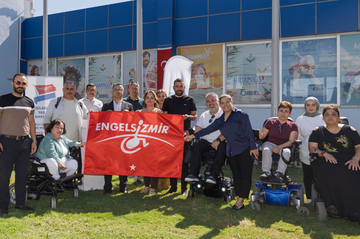 Bir grup kişi, dış mekânda büyük camlı bir yapının önünde bir araya gelmiş şekilde poz veriyor. Aralarında tekerlekli sandalye kullanan bireyler de bulunuyor. Grup, üzerinde “Engelsiz İzmir” yazan kırmızı bir bayrak tutuyor. Yan tarafta dikey bir bilgilendirme afişi yer alıyor. Arka plandaki binanın camlarında “Aquapolis” yazılı afişler görülüyor. Sahne, erişilebilirlik ve engelli haklarına yönelik bir etkinlik atmosferi taşıyor.