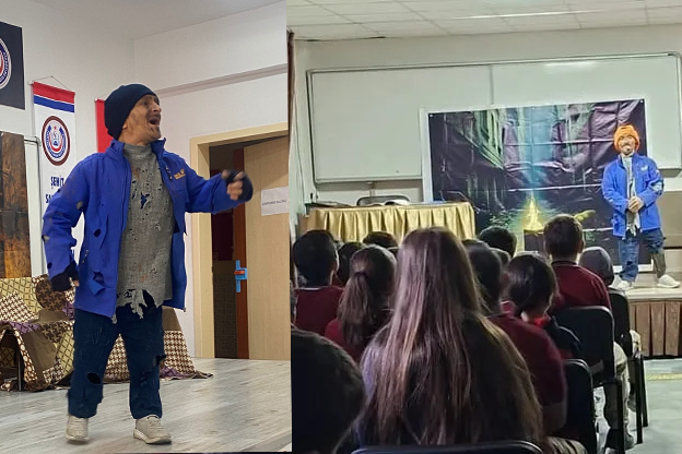 Yan yana yerleştirilmiş iki sahne görülüyor. Solda, bir oyuncu kapalı bir salonda ahşap zeminde duruyor; mavi ceket, bere ve spor ayakkabılarla bir performans sergiliyor. Arkada duvarda bayraklar ve bir kapı bulunuyor. Sağda aynı oyuncu küçük bir sahnede, kırmızı üniformalı çocuklardan oluşan bir izleyici grubuna hitap ediyor. Sahne arkasında masalsı bir sokak dekoru yer alıyor.