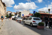 Görselin üst bölümünde, şehir merkezinde yer alan açık bir sokak ve otopark alanı yer alıyor. Yolun sağ kenarında, üzerinde tekerlekli sandalye simgesi bulunan mavi engelli park tabelasının hemen yanına usulsüz şekilde park etmiş beyaz renkli bir binek otomobil görülüyor; aracın önünde saksı içinde yeşil bir süs bitkisi, gerisinde ise duba ve koniler yer alıyor. Kaldırımda yürüyen yayalar, arka planda gölgelikli tezgâhlar ve dükkânlar ile güneşli, parçalı bulutlu gökyüzü altında uzanan şehir binaları seçiliyor.