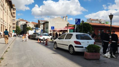 Görselin üst bölümünde, şehir merkezinde yer alan açık bir sokak ve otopark alanı yer alıyor. Yolun sağ kenarında, üzerinde tekerlekli sandalye simgesi bulunan mavi engelli park tabelasının hemen yanına usulsüz şekilde park etmiş beyaz renkli bir binek otomobil görülüyor; aracın önünde saksı içinde yeşil bir süs bitkisi, gerisinde ise duba ve koniler yer alıyor. Kaldırımda yürüyen yayalar, arka planda gölgelikli tezgâhlar ve dükkânlar ile güneşli, parçalı bulutlu gökyüzü altında uzanan şehir binaları seçiliyor.